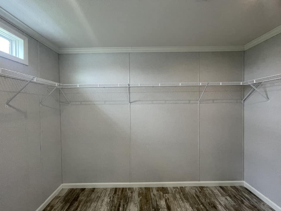 Empty walk-in closet with light gray walls, hardwood flooring, and a small window. Two metal shelves are mounted on adjacent walls. Bright, minimalistic.