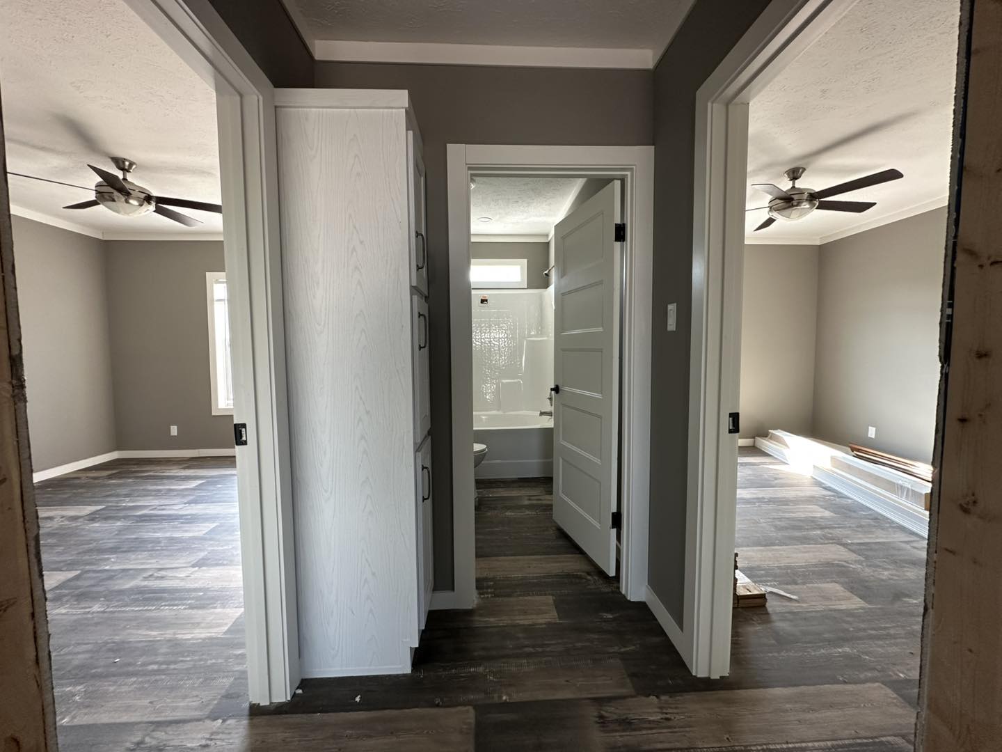 View of a hallway in a modern home. The hallway has a bathroom with a white door and shower straight ahead, and two rooms on the sides with ceiling fans and wood flooring. The color scheme is neutral, with a calm, clean, and minimalistic tone.