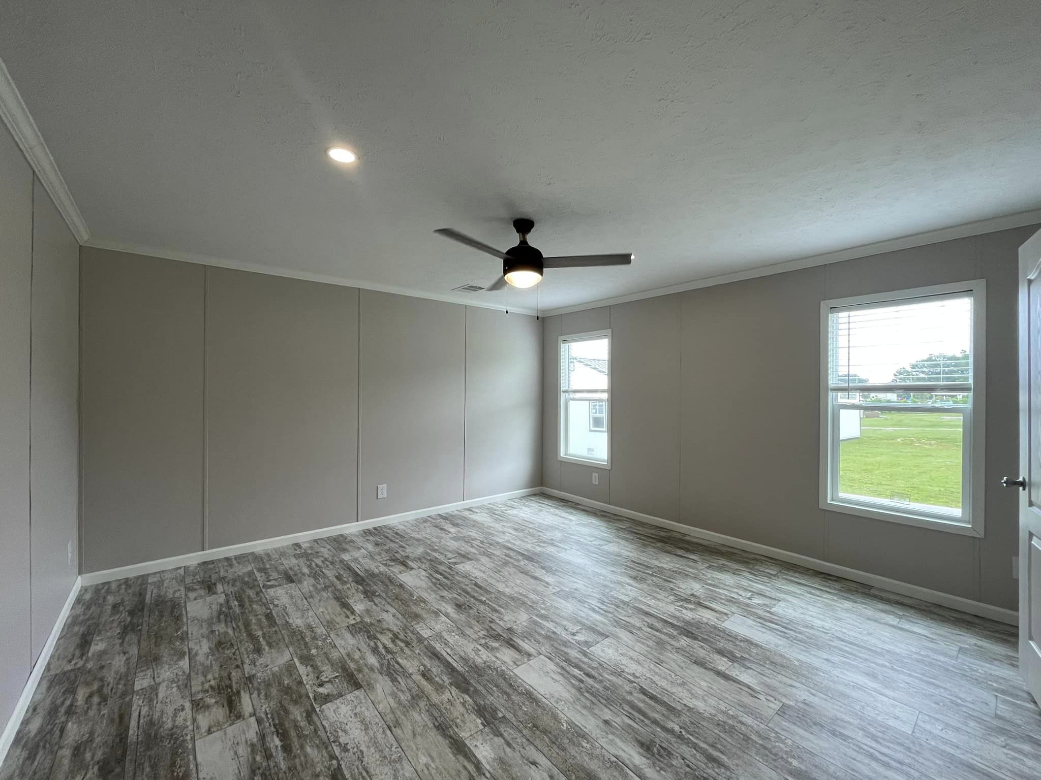 Spacious empty room with light gray walls and wood-look flooring. Two windows allow natural light, and a ceiling fan hangs centrally, creating a modern ambiance.