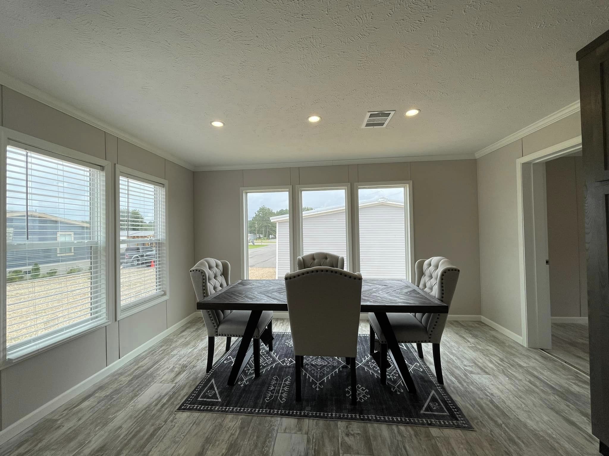 Bright dining room with a wooden table and four tufted chairs on a patterned rug. Large windows offer natural light and a view outside.