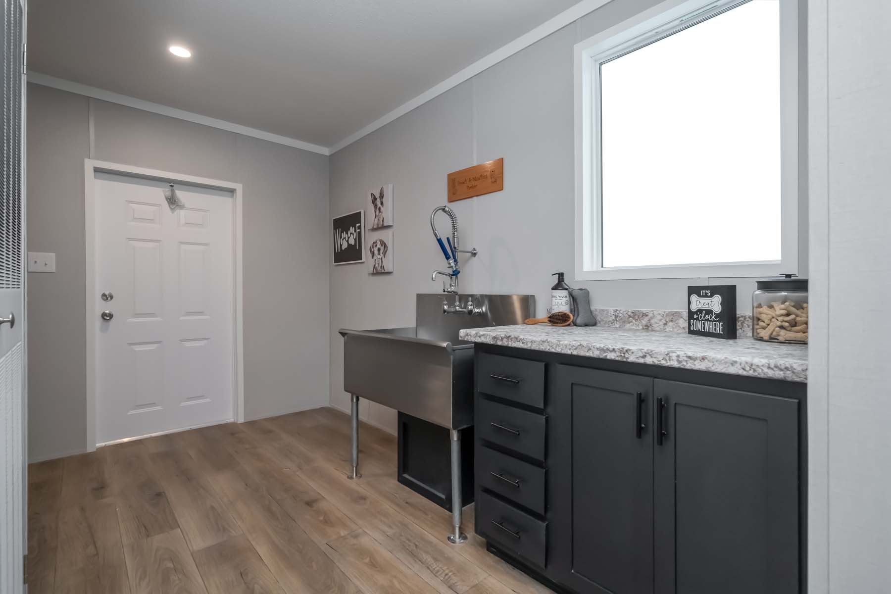 A clean dog washing room with a stainless steel sink, black cabinetry, and marble countertop. Wall decor includes dog-themed art and a bright window.