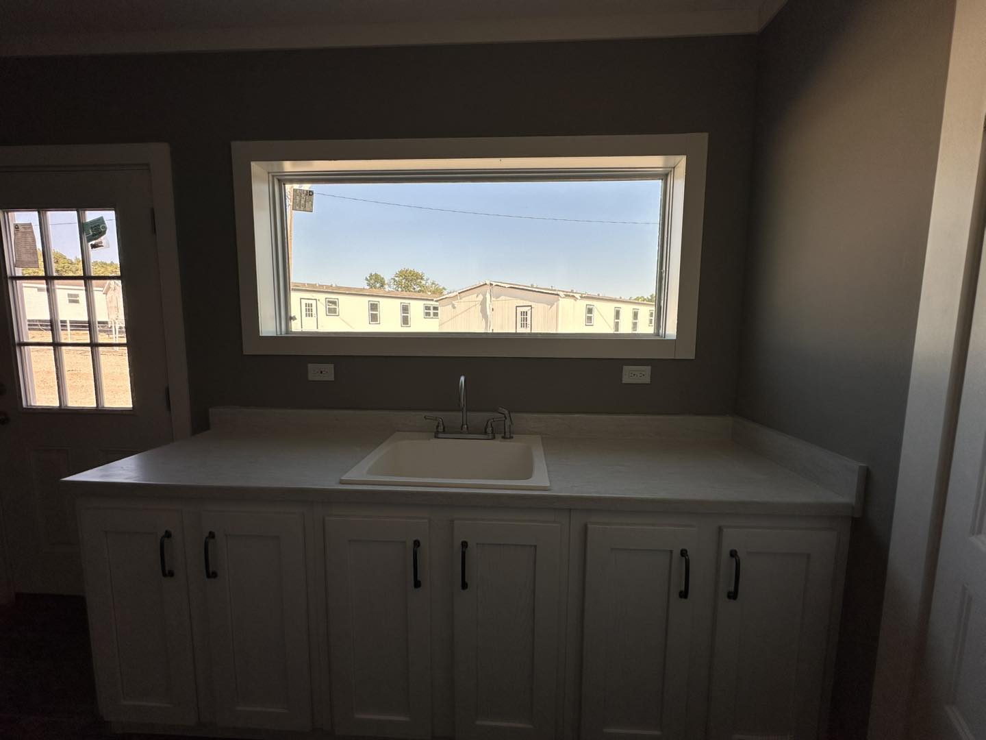 Modern kitchen sink with white countertop under a wide window, showing a view of buildings and trees outside. The room has a calm, neutral tone.