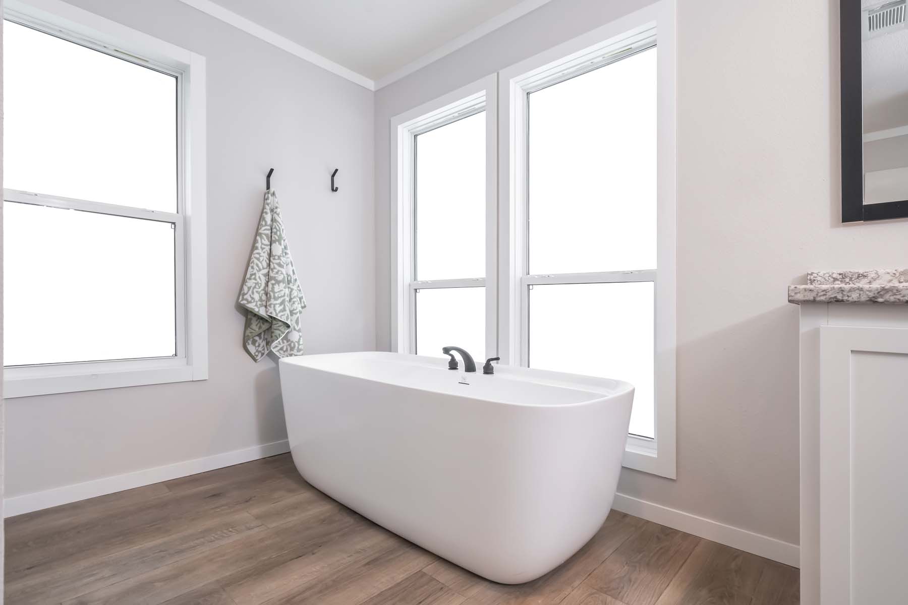 Minimalist bathroom with a freestanding white tub by two large windows, a patterned towel on a hook, and wood flooring, conveying calmness.