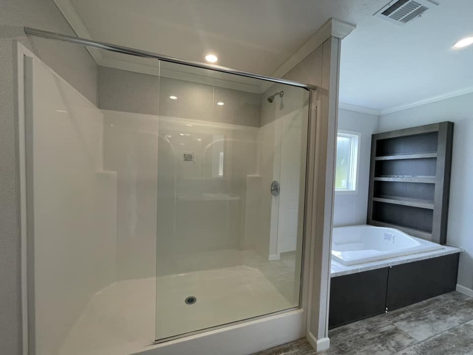 Modern bathroom with a large glass-enclosed shower and a sunlit corner bathtub beside a wooden shelving unit. The room is sleek and minimalist.