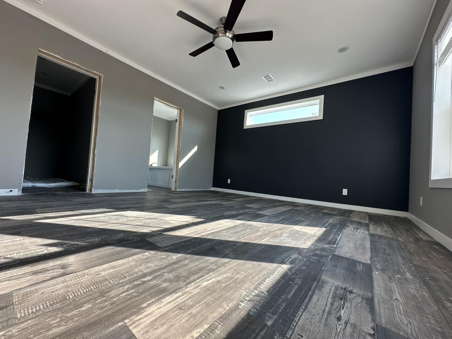 A modern room featuring dark wood flooring, gray walls, and a black accent wall. A ceiling fan and large window create a bright, spacious feel.