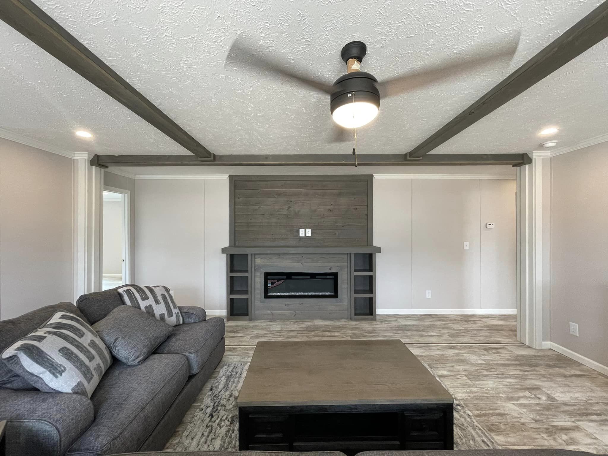 Modern living room with gray sofa, geometric-patterned pillows, a dark wooden coffee table, and a wall-mounted electric fireplace. Ceiling fan above.
