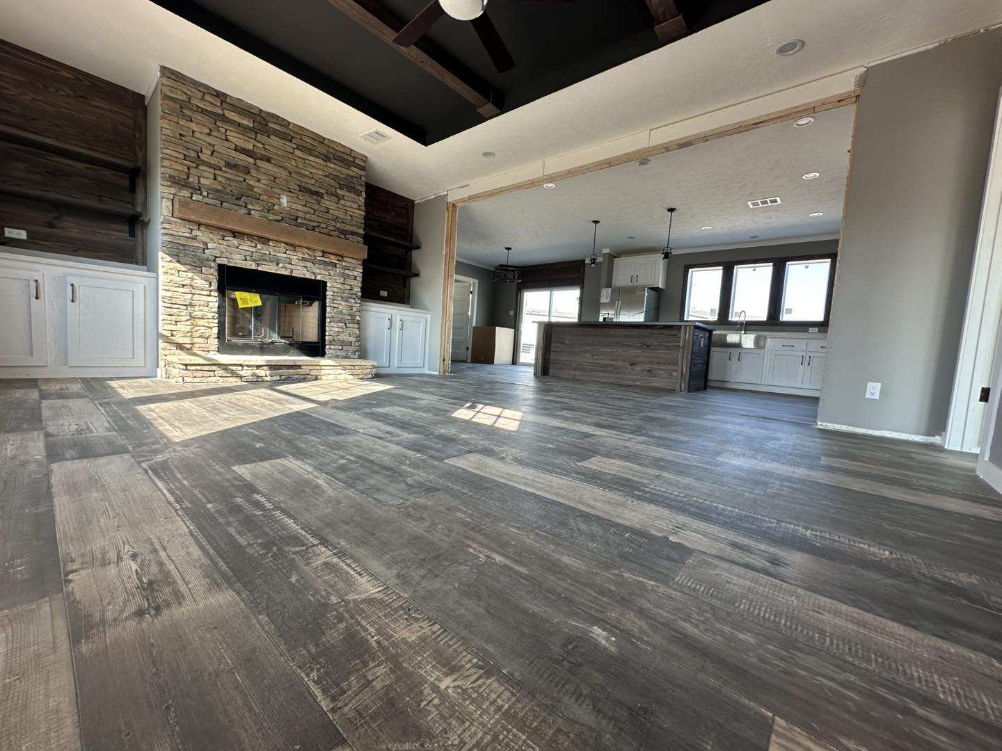 Spacious living area with dark wood flooring, stone fireplace, and modern open kitchen. Sunlight streams through large windows, creating a warm, inviting atmosphere.