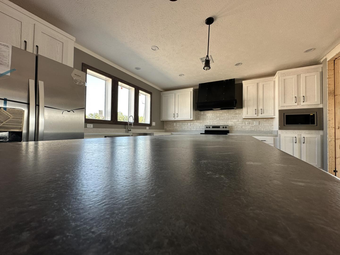 Modern kitchen with a spacious, dark countertop in the foreground. Features include white cabinets, stainless steel appliances, and large windows. Bright and inviting.