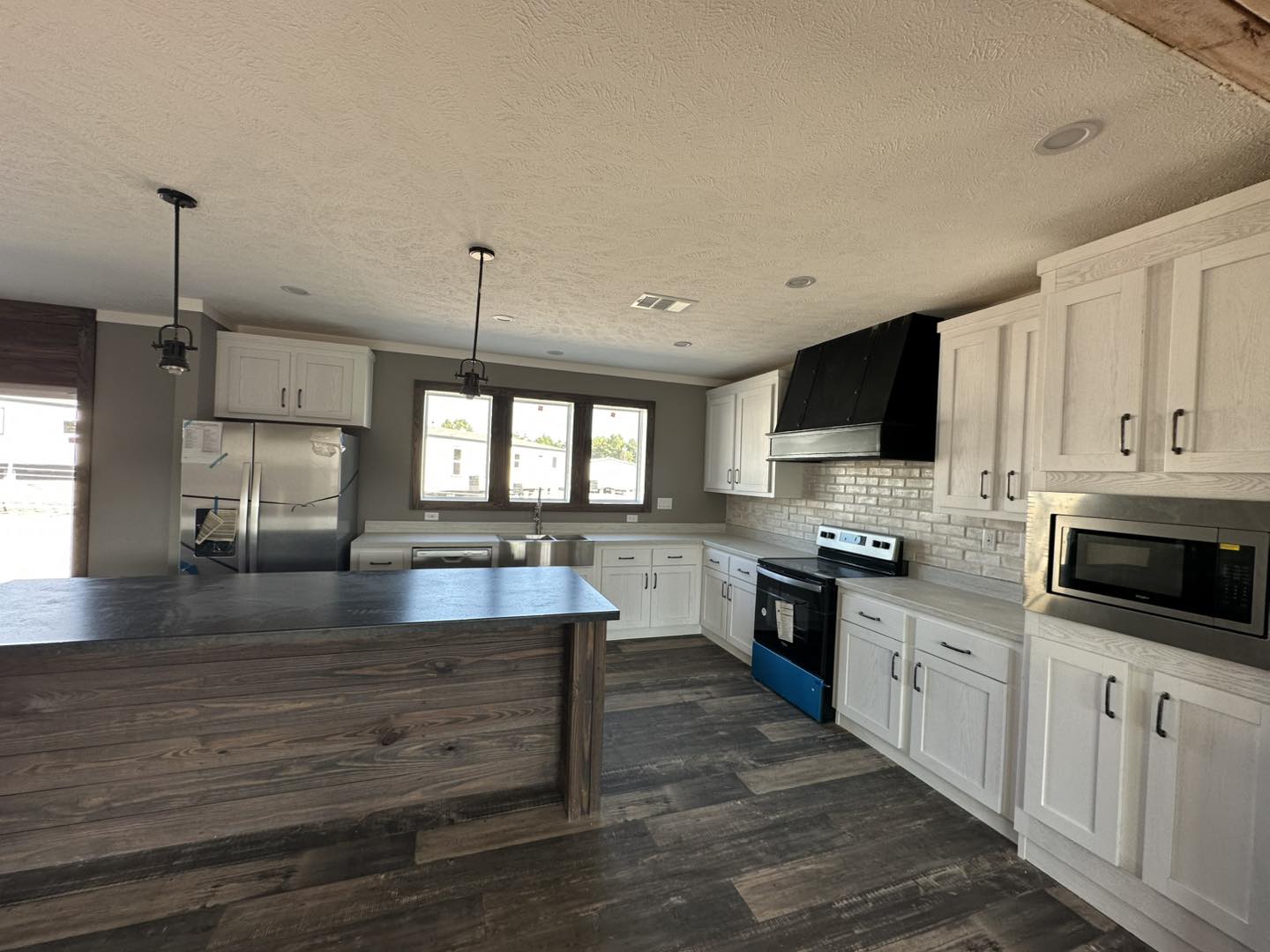 Modern kitchen with dark wood flooring, white cabinets, and stainless steel appliances. Two pendant lights hang over a wood island. Bright and spacious.