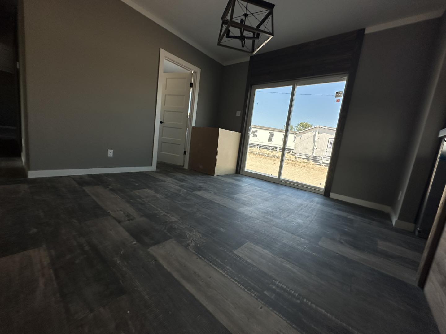 Modern room with dark wood flooring, gray walls, and a geometric ceiling light. A door and large window show a clear, sunny view outside.