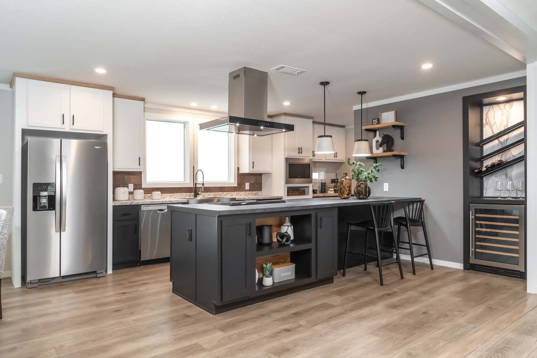 Modern kitchen with light wood floors, a central island with black cabinets, stainless steel appliances, sleek lighting, and a wine rack on the wall.