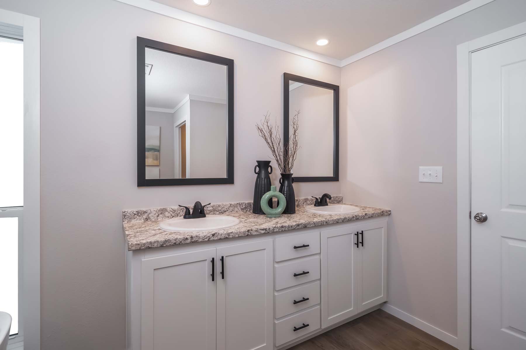 Modern bathroom with dual sinks on a speckled marble counter. Black-framed mirrors hang above. Decor includes vases and twigs, creating a calm ambiance.