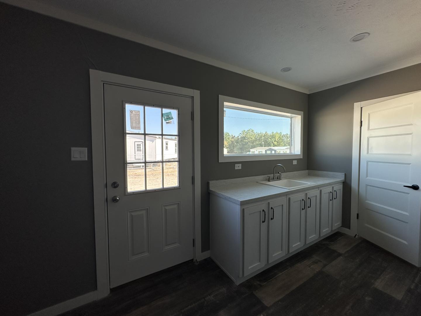 A small, modern laundry room with gray walls and wood floor. It features a white cabinet with a sink under a wide window, adjacent to a white door.