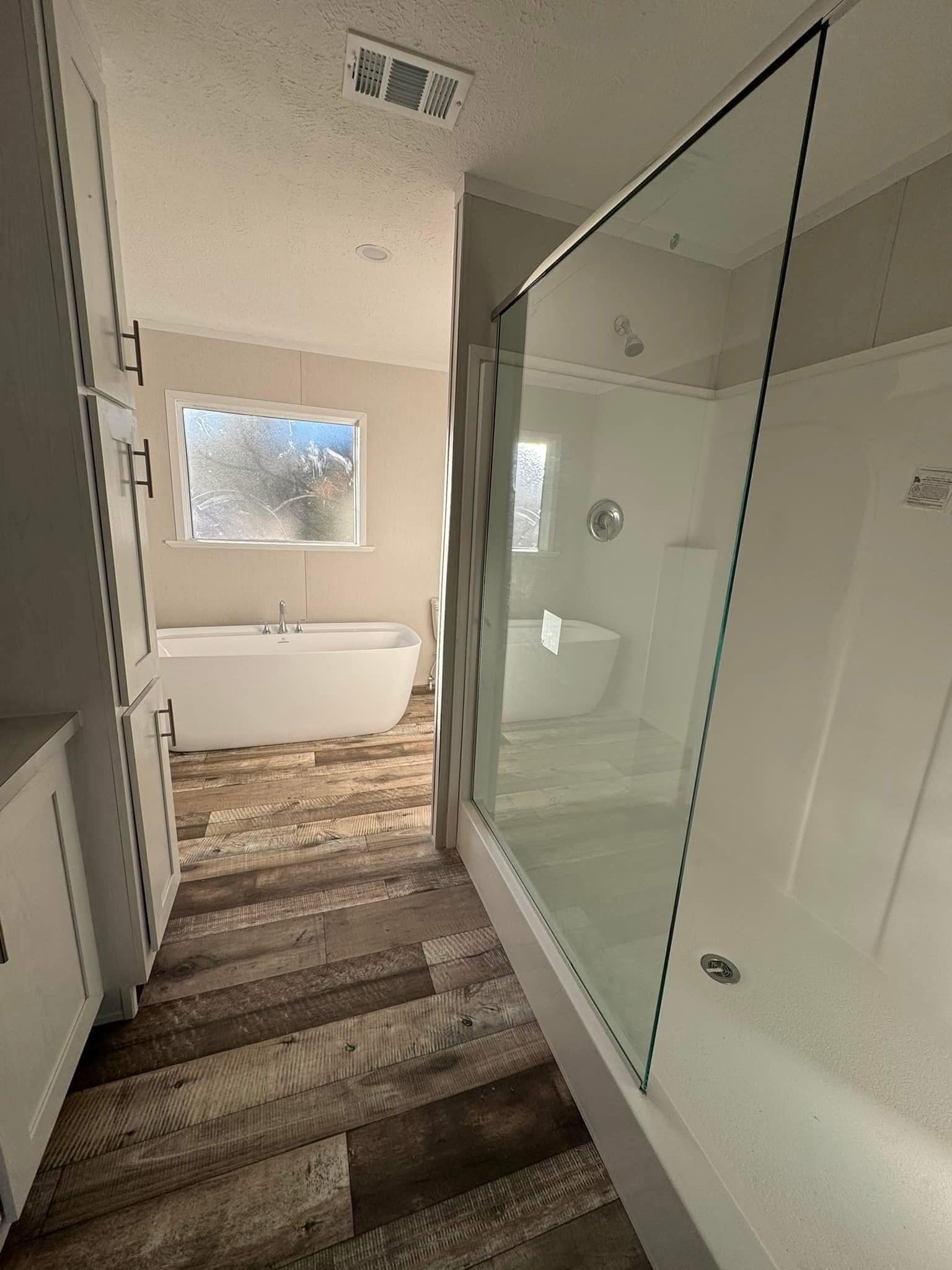 Modern bathroom with a freestanding tub by a frosted window, glass-enclosed shower, and wood plank flooring. Bright, serene ambiance.