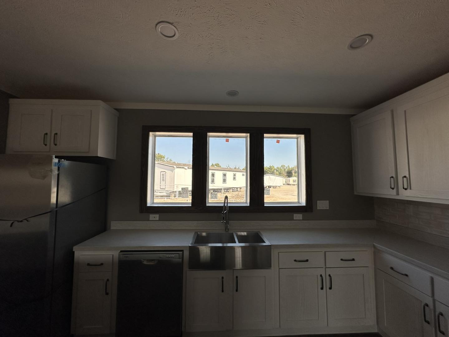 Modern kitchen with stainless steel appliances and white cabinets. Large window over sink reveals a clear, sunny view of buildings and greenery outside.