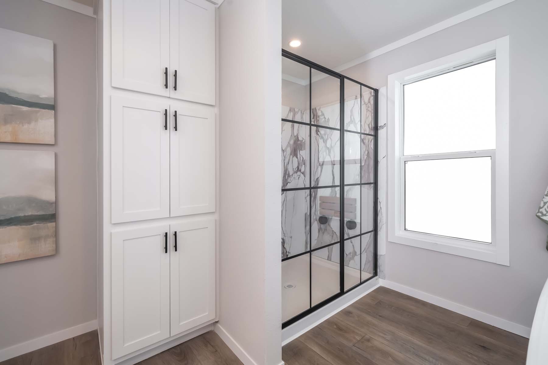 Modern bathroom with a black-framed glass shower and marble-patterned tiles. Beside the shower is a tall white cabinet, and the room has a light, airy feel.