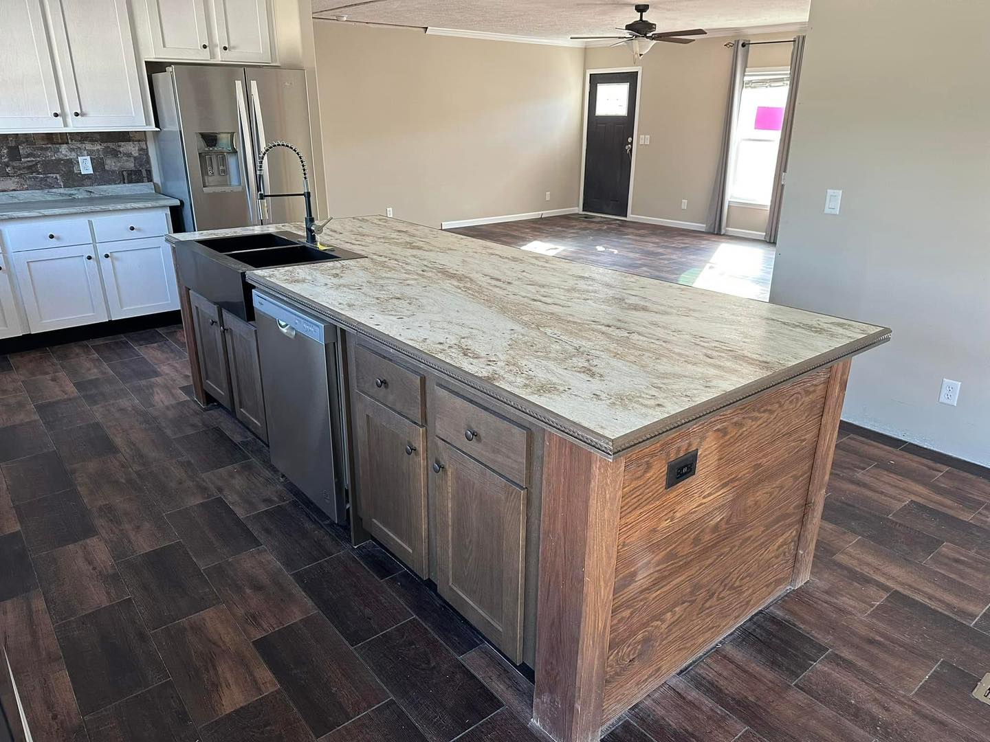 Spacious kitchen with a large, beige marble island and wood cabinets. Dark wood flooring, stainless steel fridge, sink, and dishwasher. Bright, modern feel.