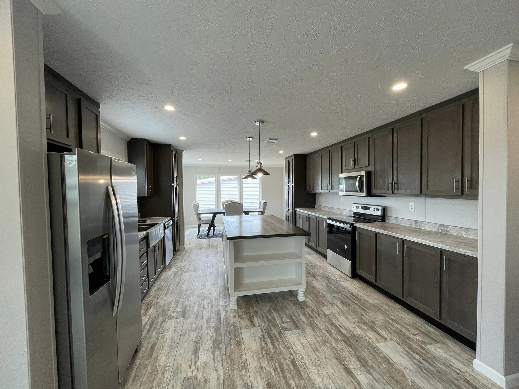 Modern kitchen with dark wooden cabinets, stainless steel appliances, and a central island. Bright lighting and a dining area with windows in the background.
