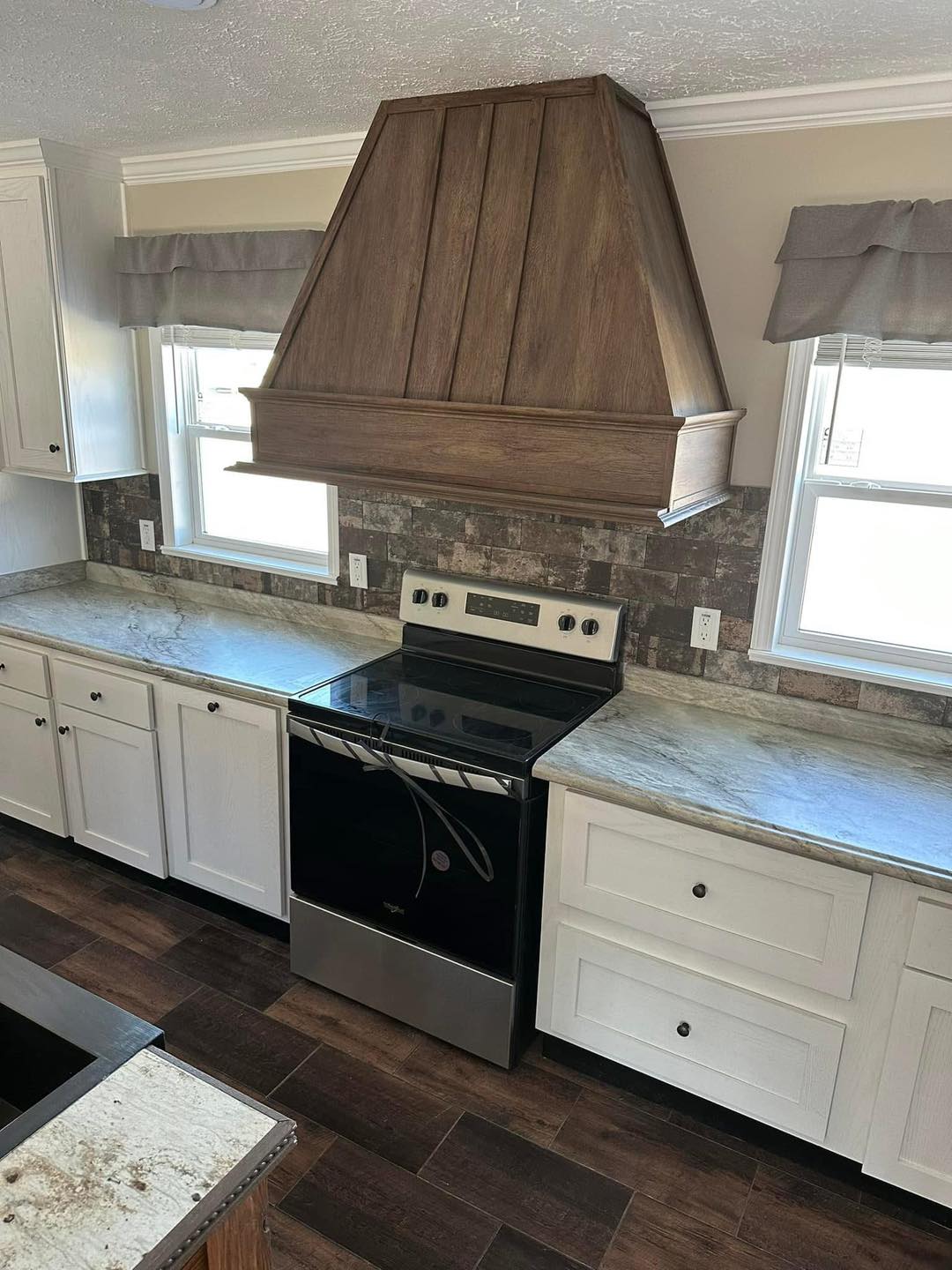 A modern kitchen with a black electric stove beneath a wooden range hood. White cabinets and marble countertops enhance the bright, airy atmosphere.