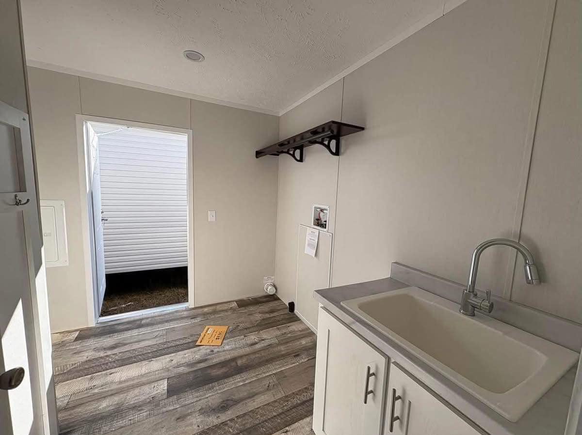 Small, empty laundry room with light gray walls and wood-look vinyl flooring. It features a wooden shelf, sink with cabinet, and an open door.