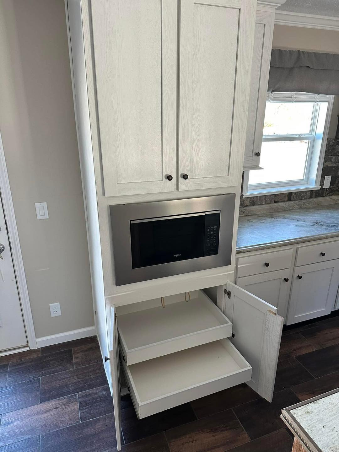Modern kitchen with white cabinets, featuring a built-in microwave and two open pull-out shelves below. Sunlit window and dark wood flooring create a warm ambiance.
