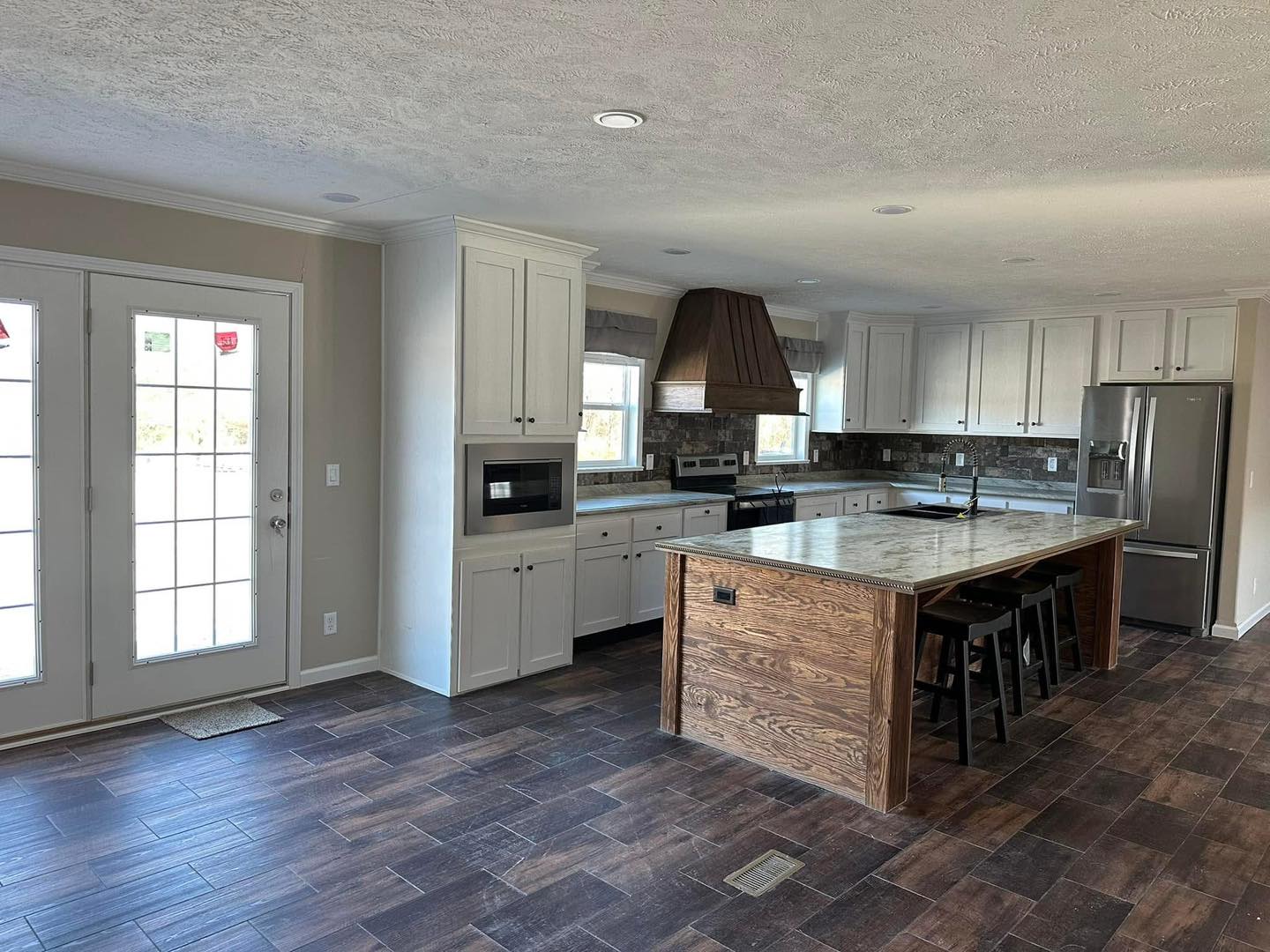 Spacious kitchen with dark wooden floors, white cabinets, and a central island with a marble countertop. A stainless steel fridge and range hood sit in the background. French doors on the left allow natural light.