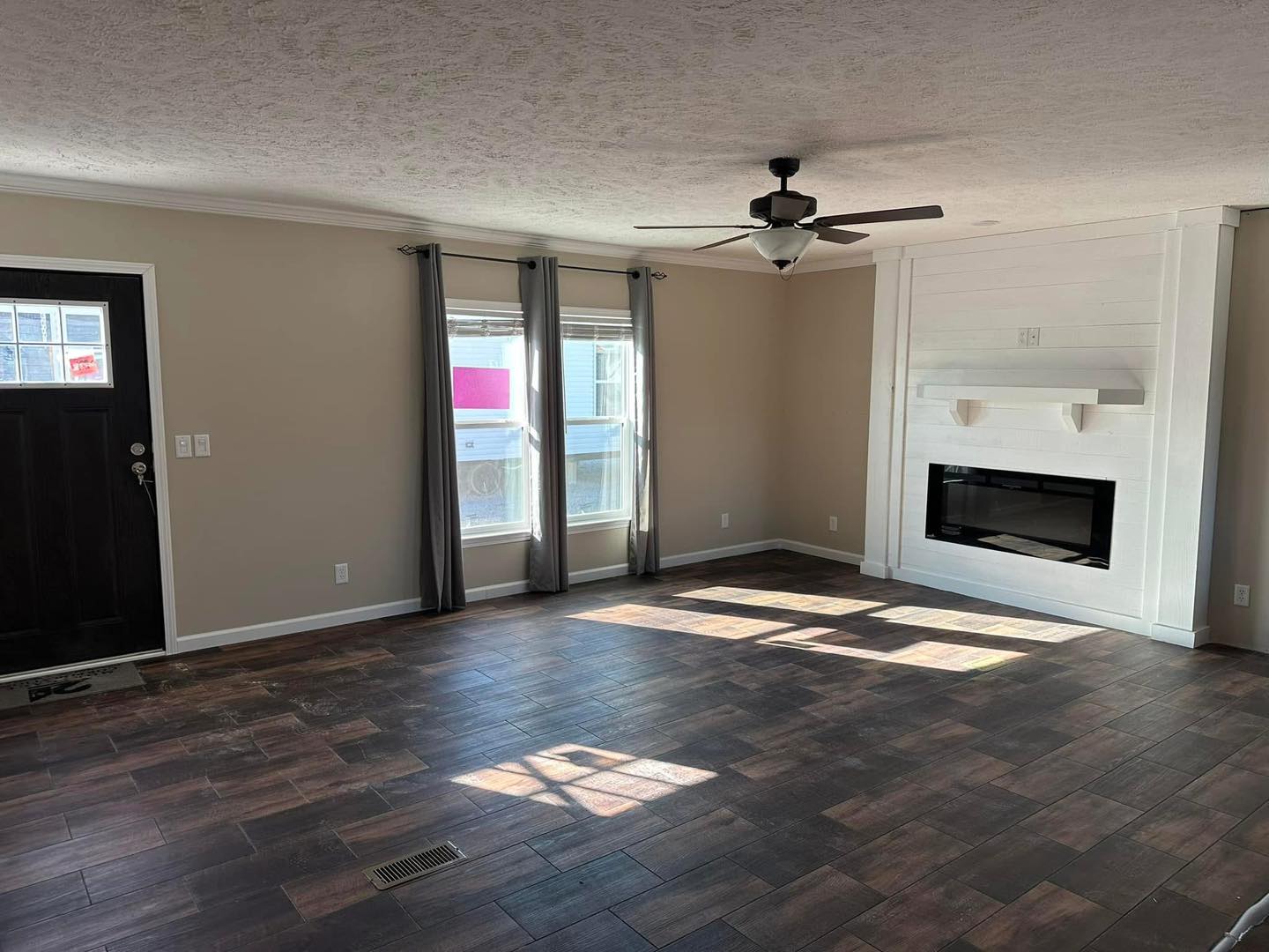 A spacious living room with dark wood flooring and beige walls. Features a black ceiling fan, large windows with gray curtains, and a white fireplace.