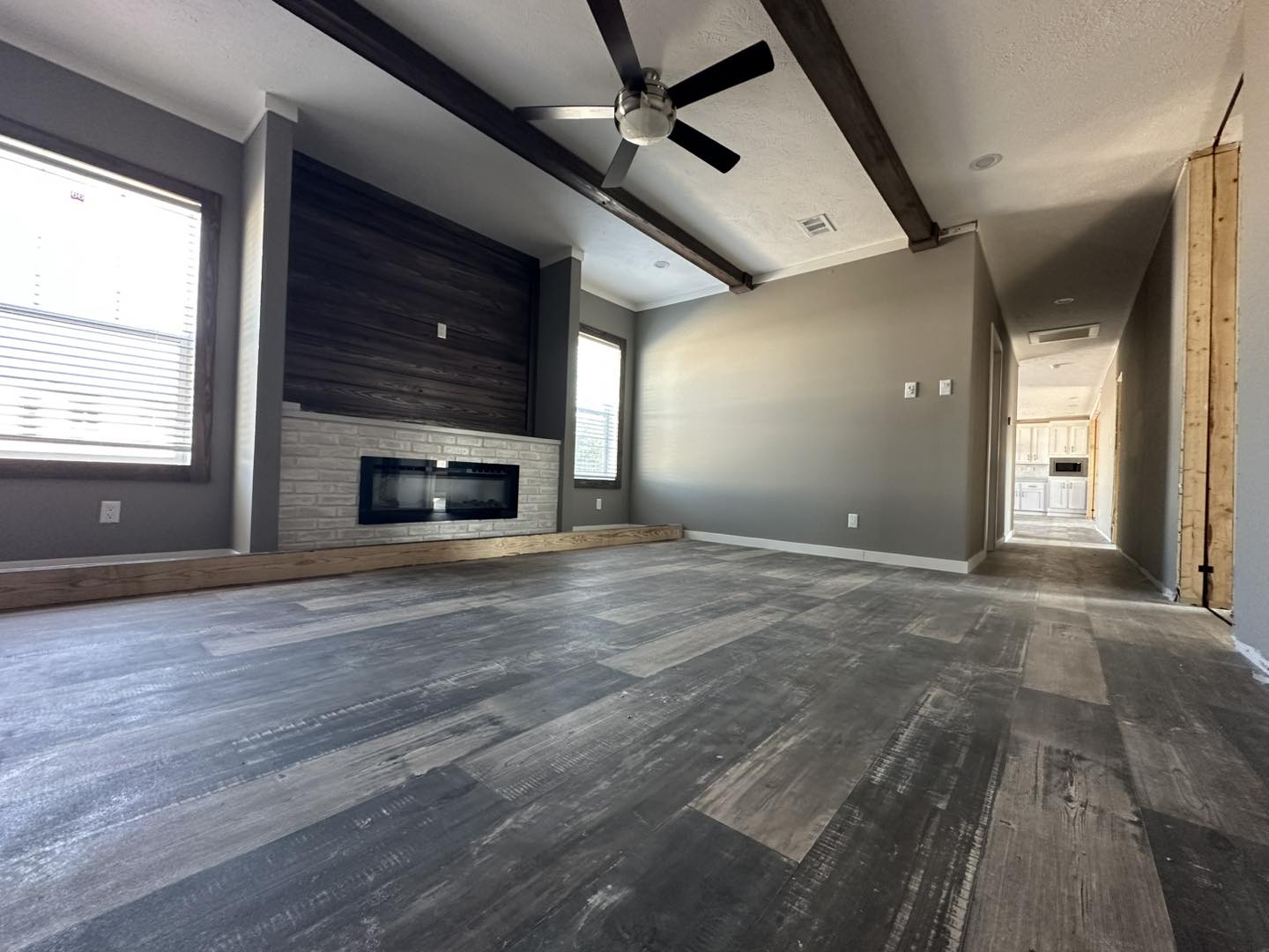 Modern living room with rustic wood flooring and ceiling beams, featuring a sleek electric fireplace against a dark accent wall. Soft natural light enters through two large windows.