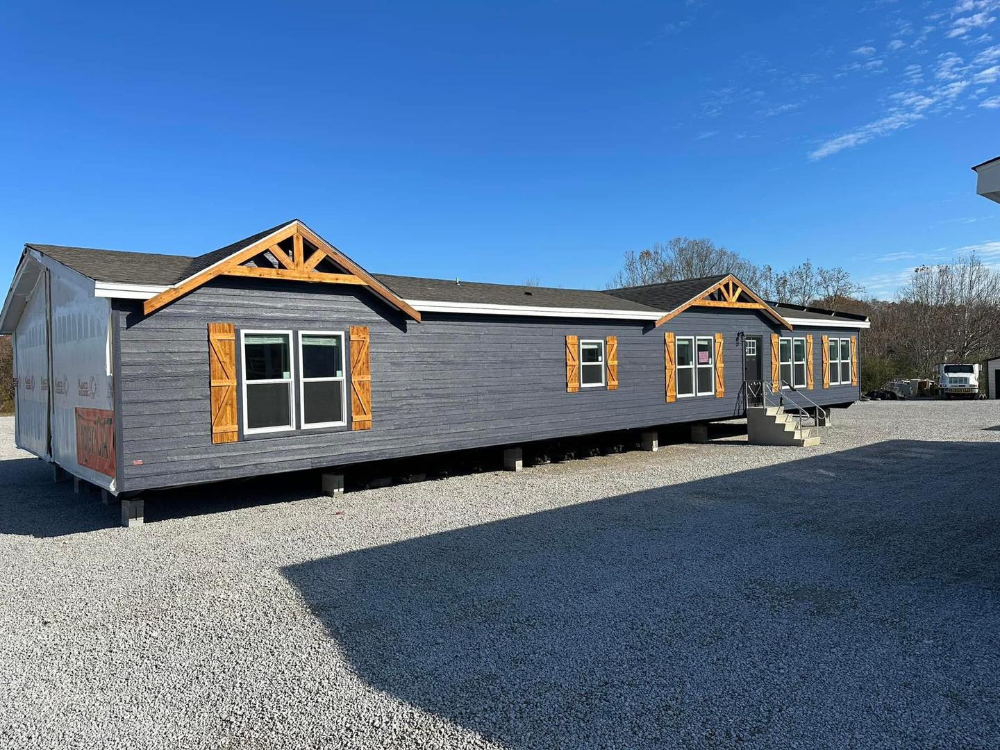 A long, gray manufactured home with wooden shutters and triangular trim sits on a gravel lot under a clear blue sky, conveying a sense of simplicity and functionality.
