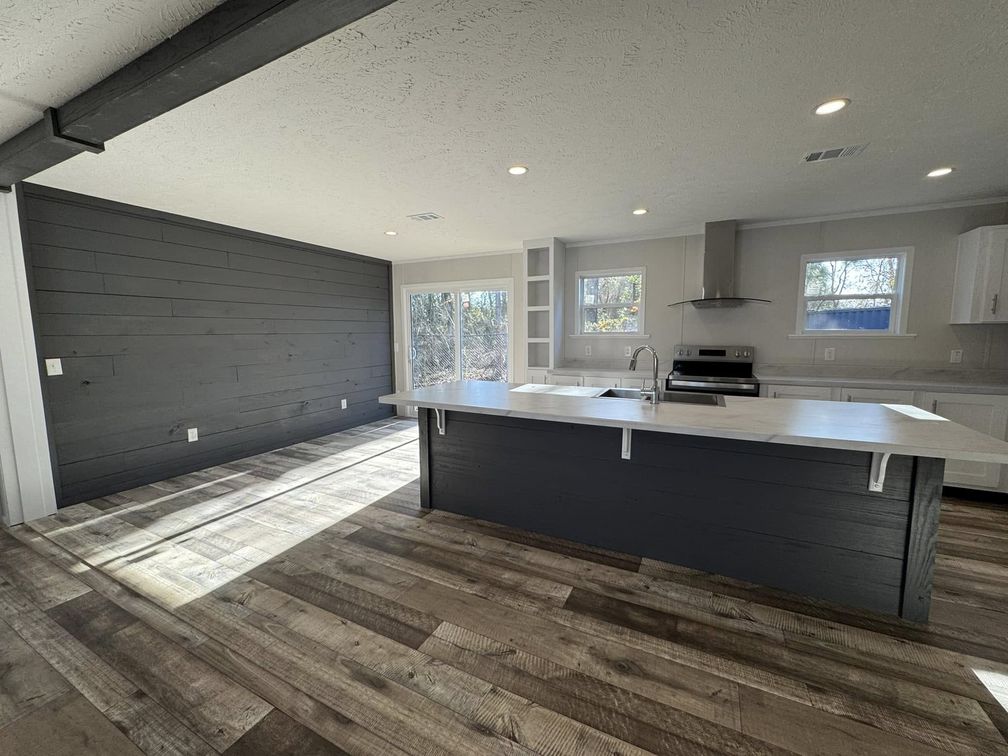 Modern kitchen with dark wood floors and a black accent wall. Light gray island, cabinets, stainless steel appliances, and large windows create an open, airy feel.
