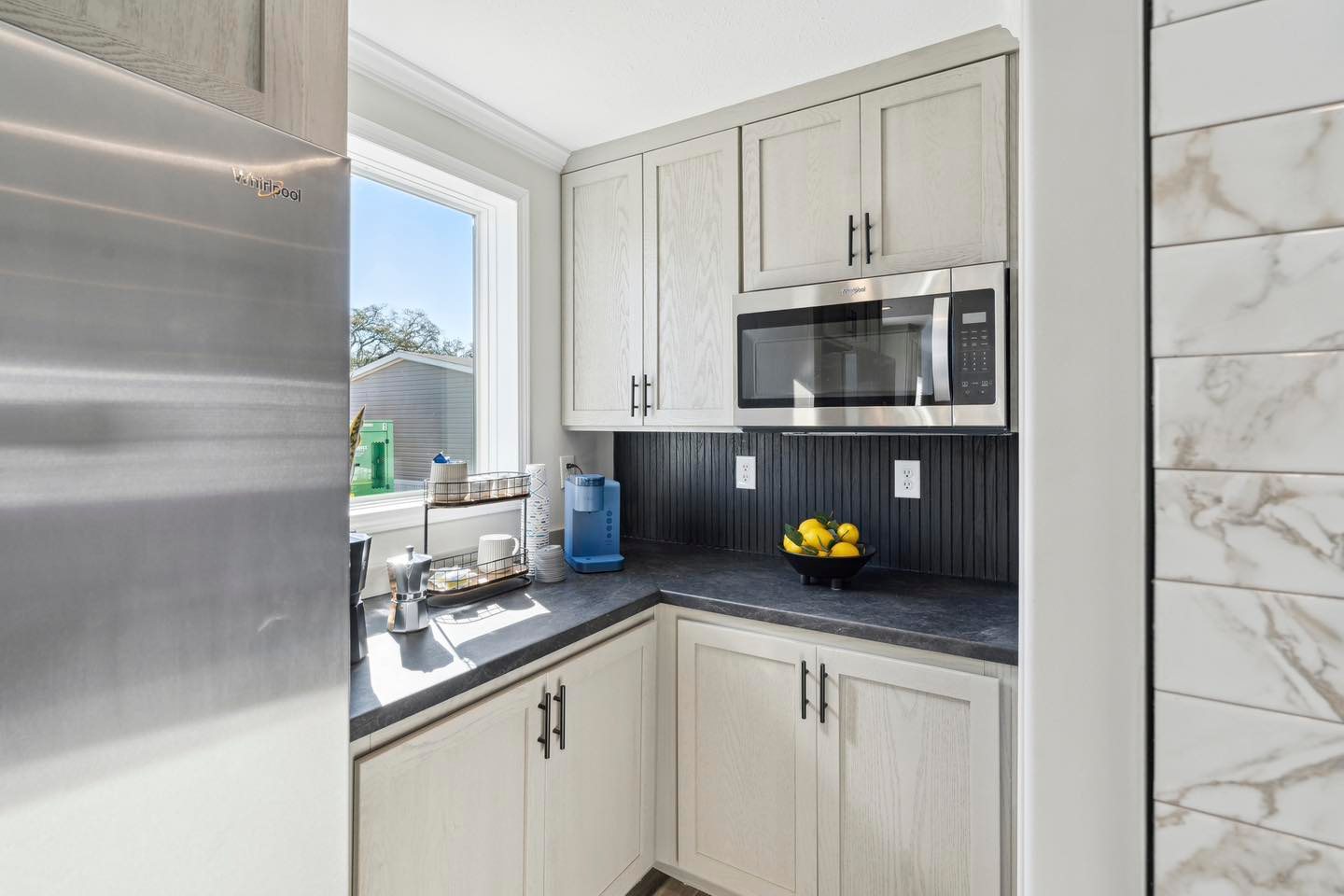 Compact kitchen with light wood cabinets, dark countertops, and stainless steel appliances. A bowl of lemons adds color, while sunlight streams through a window.