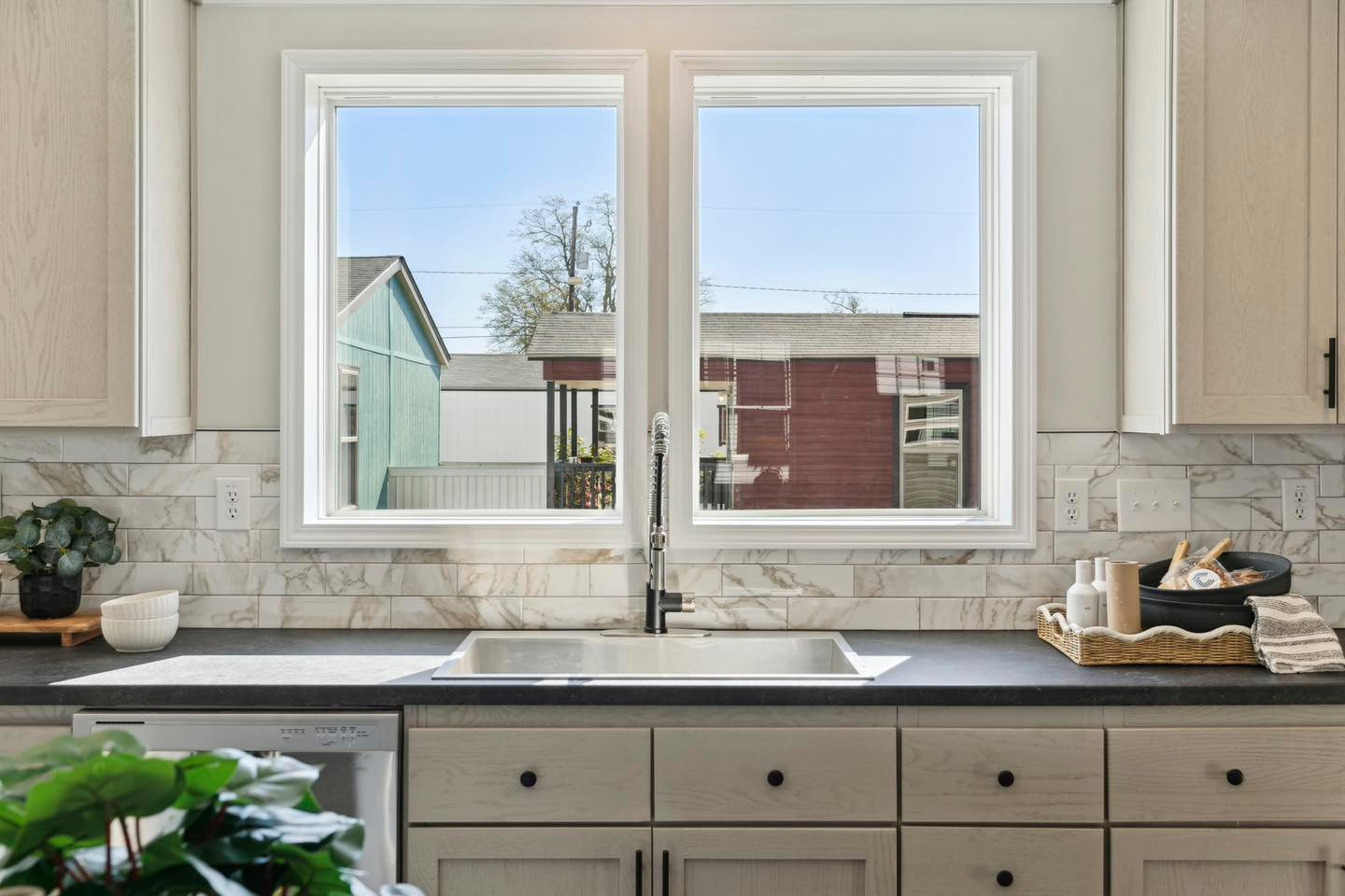 A kitchen sink is centered below two large windows, framed by light wood cabinets. Sunlight streams in, illuminating the marble backsplash and countertop decor.