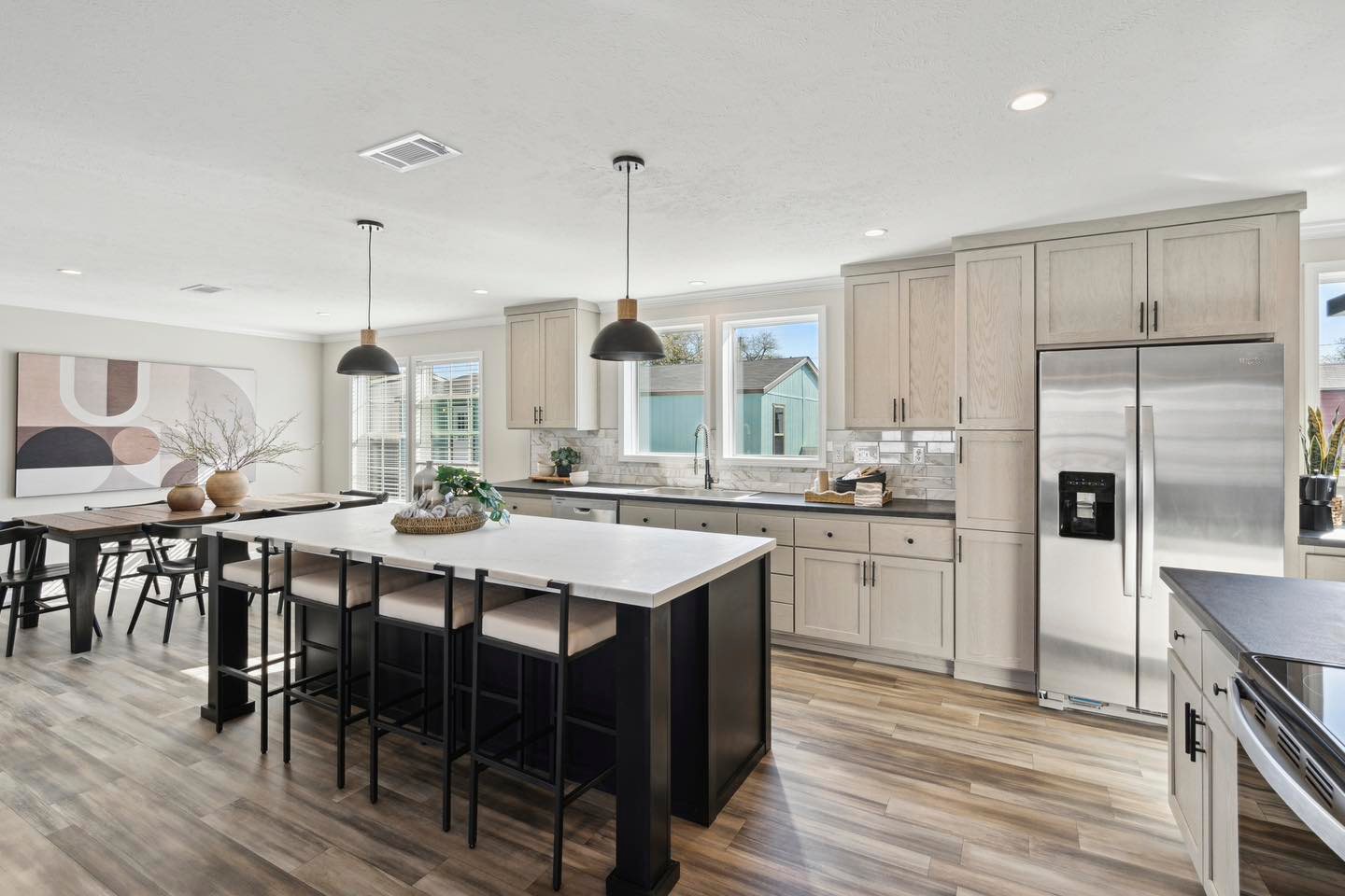 Modern kitchen with light wood cabinets, a large island with seating, and pendant lights. Dining area features abstract art and natural decor. Bright and spacious.