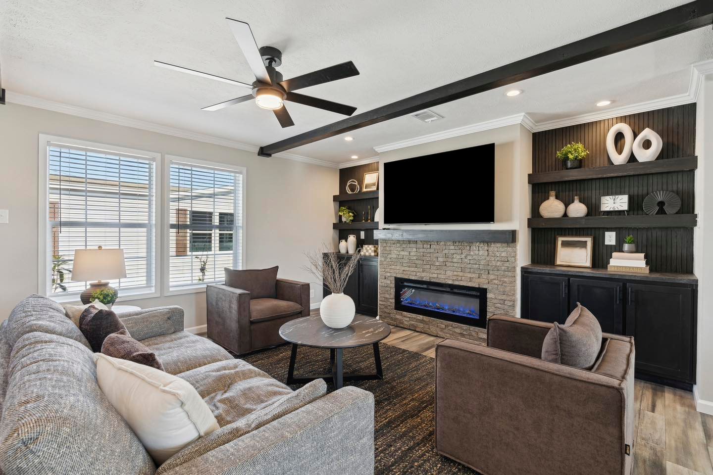 A modern living room with a gray sofa, two brown armchairs, and a round coffee table. Features a stone fireplace, large TV, shelves with decor, and natural light from large windows.