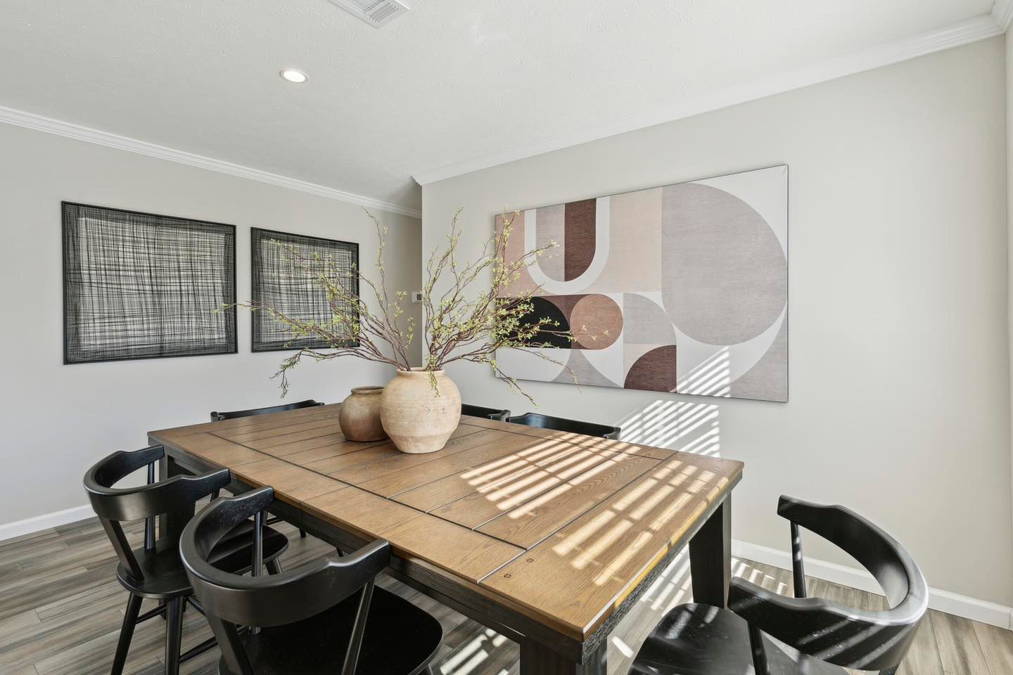 A modern dining room with a wooden table surrounded by six black chairs. Two vases with branches sit on the table. Abstract art and framed grid patterns adorn the light gray walls. Sunlight streams through a window, casting geometric shadows, creating a calm, stylish ambiance.