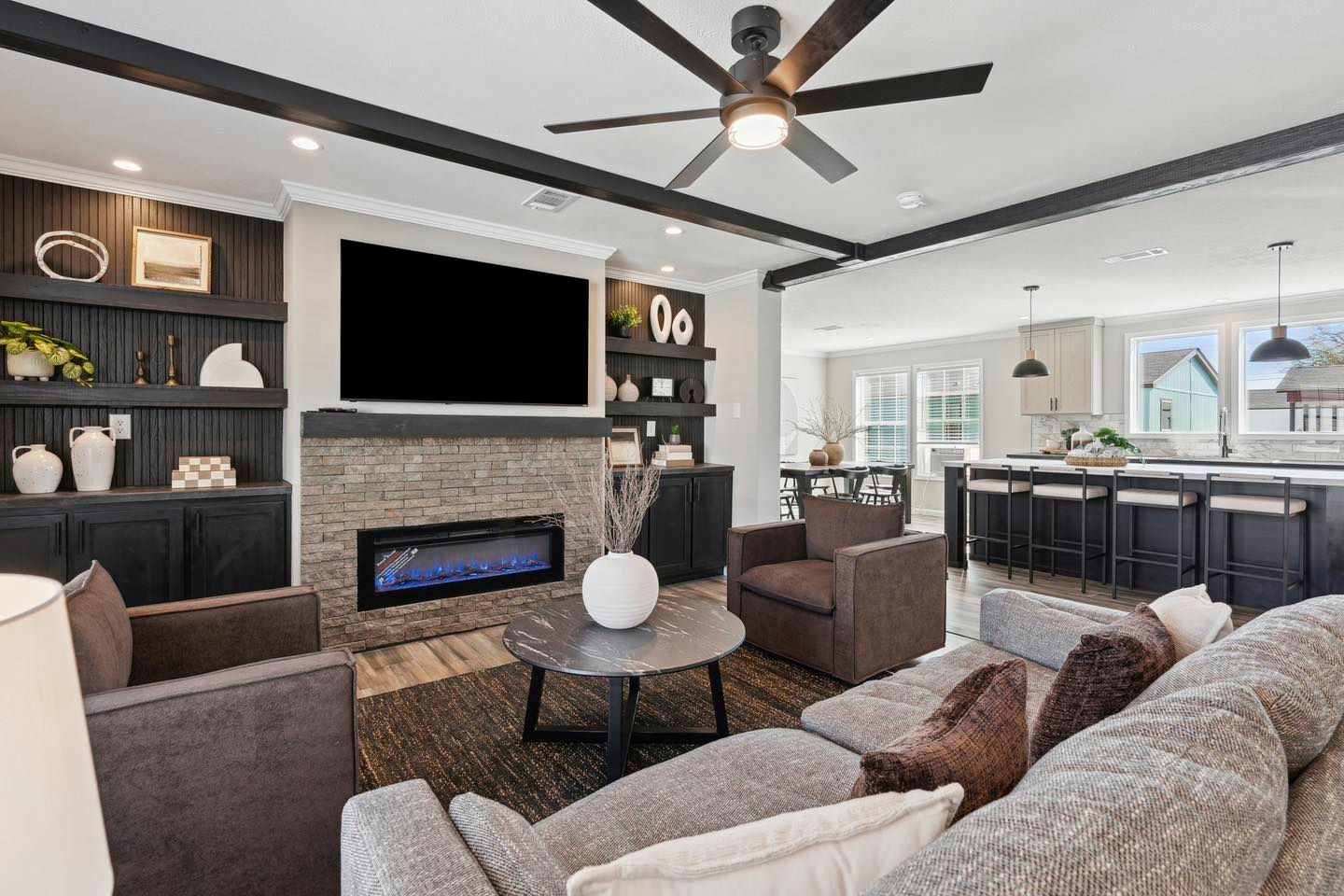 Modern living room with a gray sectional, brown armchairs, and a black round coffee table. Stone fireplace with a flat-screen TV above. Open kitchen in background. Cozy and inviting atmosphere.