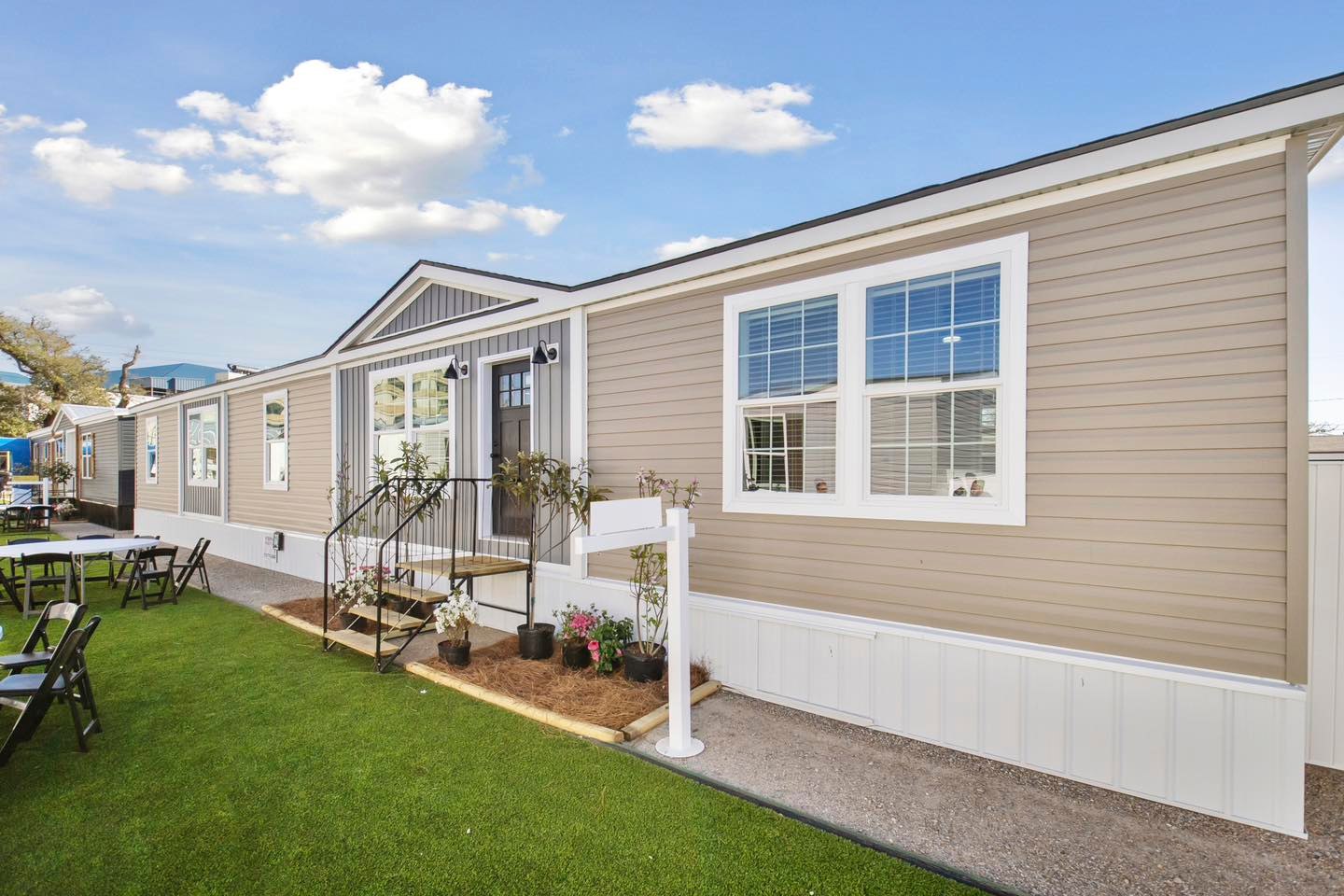 Modern beige modular home with white trim, neatly manicured lawn, potted plants by entrance, and a blue sky with fluffy clouds. Inviting and serene.