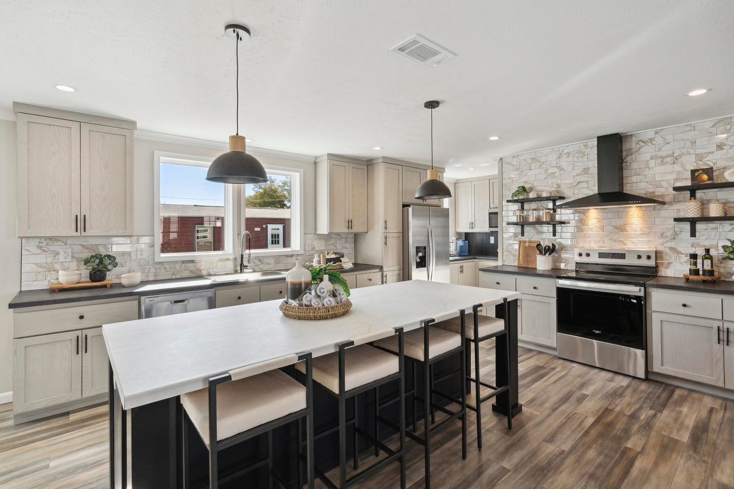 Modern kitchen with light wood cabinets, a large white island with chairs, pendant lights, and stainless steel appliances. Bright, welcoming ambiance.