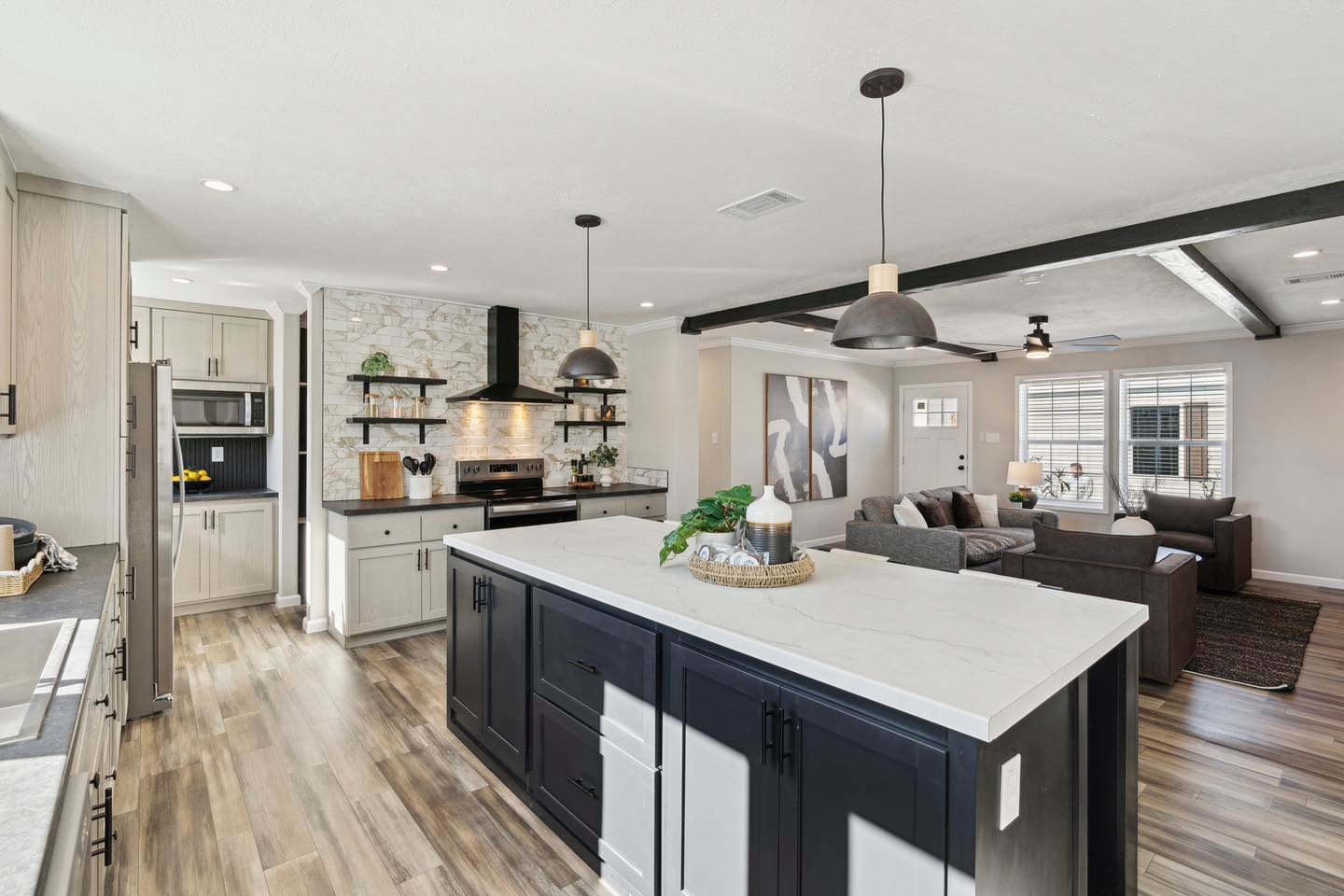 Modern open-plan kitchen and living area with light wood flooring. Features a white island, pendant lights, gray cabinetry, and a cozy gray sofa.