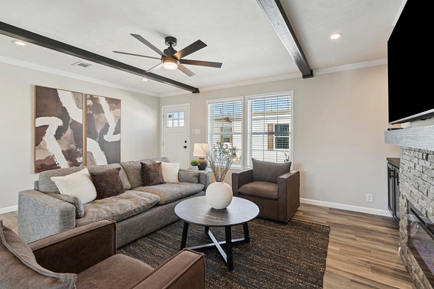 Modern living room with neutral tones featuring a gray sofa, brown armchairs, round coffee table with vase, abstract art, ceiling fan, and wide windows.