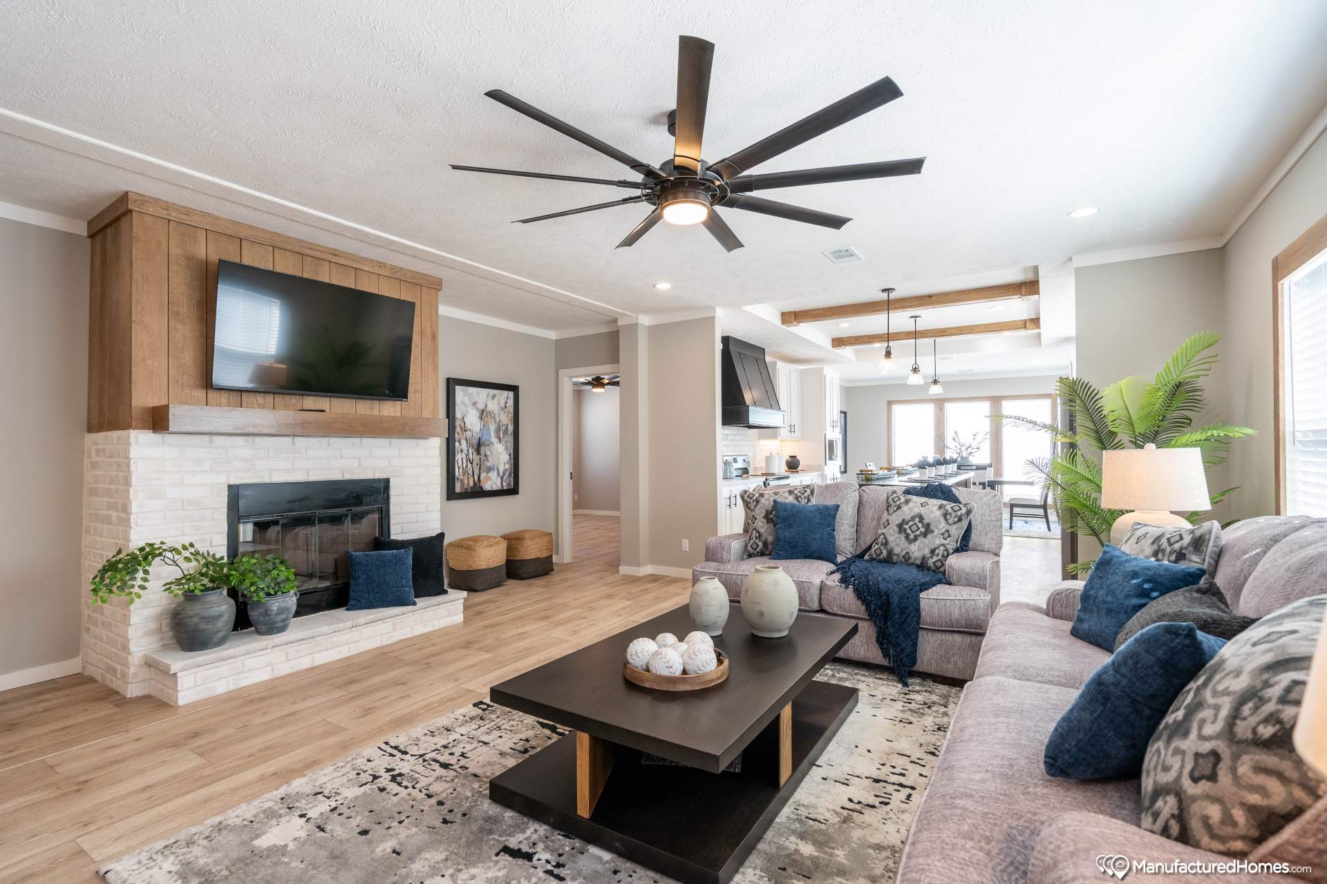 Modern living room with large ceiling fan, cozy gray sofas adorned with blue pillows, wooden coffee table, and wall-mounted TV above a white brick fireplace.