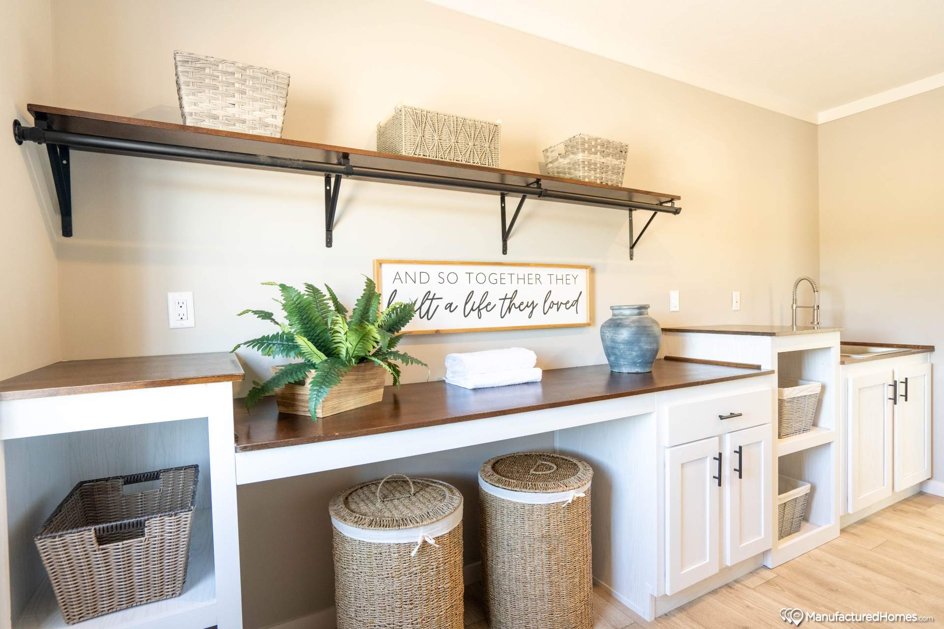 Laundry room with white cabinetry and wooden countertops. Baskets and decor, including a plant and a jar, are on the counter. A sign reads, “And so together they built a life they loved.”