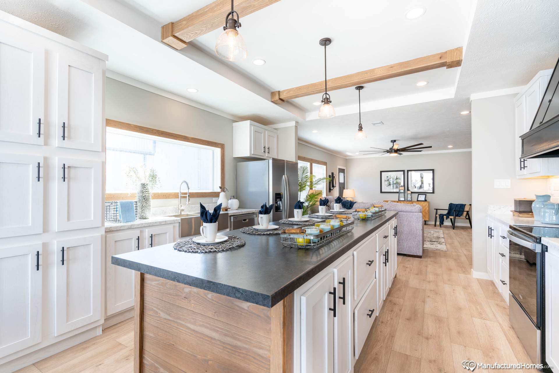 Modern kitchen and living area with white cabinets, dark countertops, and wooden beams. Bright, airy space with pendant lights and stylish decor.