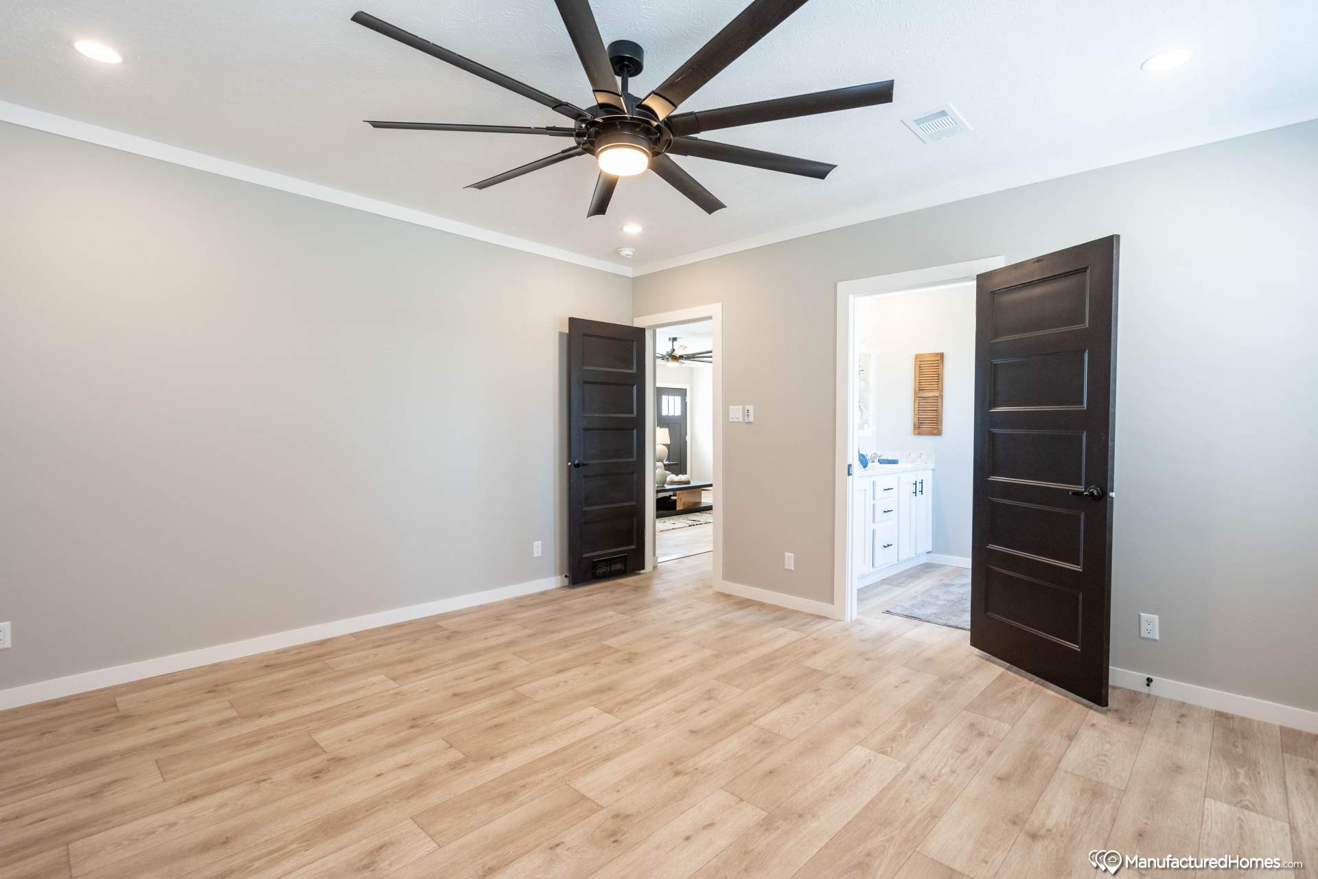 A spacious, empty room features light wood flooring and grey walls. A large ceiling fan is centered in the room. Two open dark doors reveal a bathroom and another living area, creating an airy, inviting feel.
