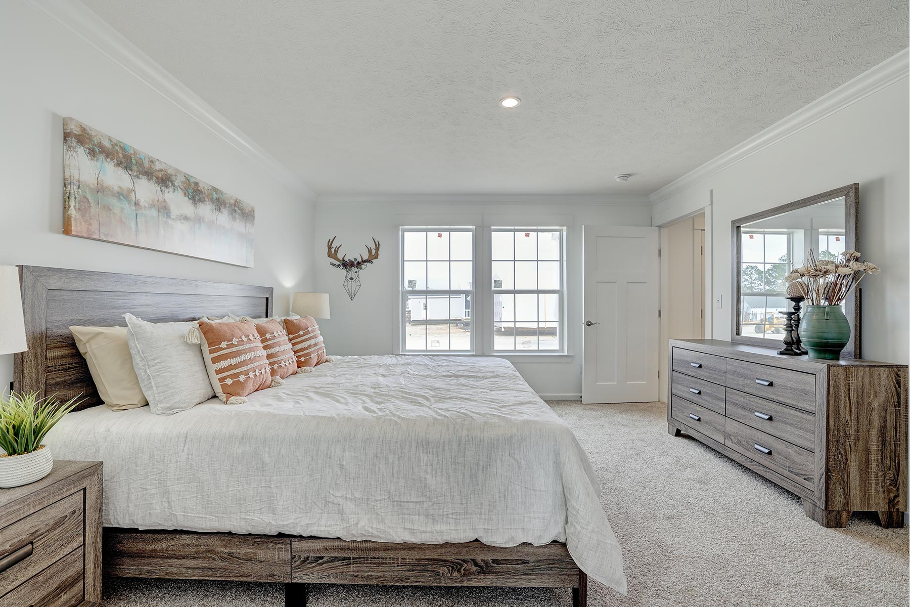 A serene bedroom with a large bed featuring white linens and decorative pillows. A wooden headboard stands against a soft gray wall. Above hangs a landscape painting. The room has a dresser with a large mirror and vase with branches. A mounted antler decor is between two large windows, letting in natural light, creating a calm and airy atmosphere.