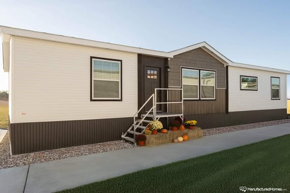 A beige and brown manufactured home with a small staircase at the entrance, decorated with pumpkins and a hay bale, set under a clear blue sky.