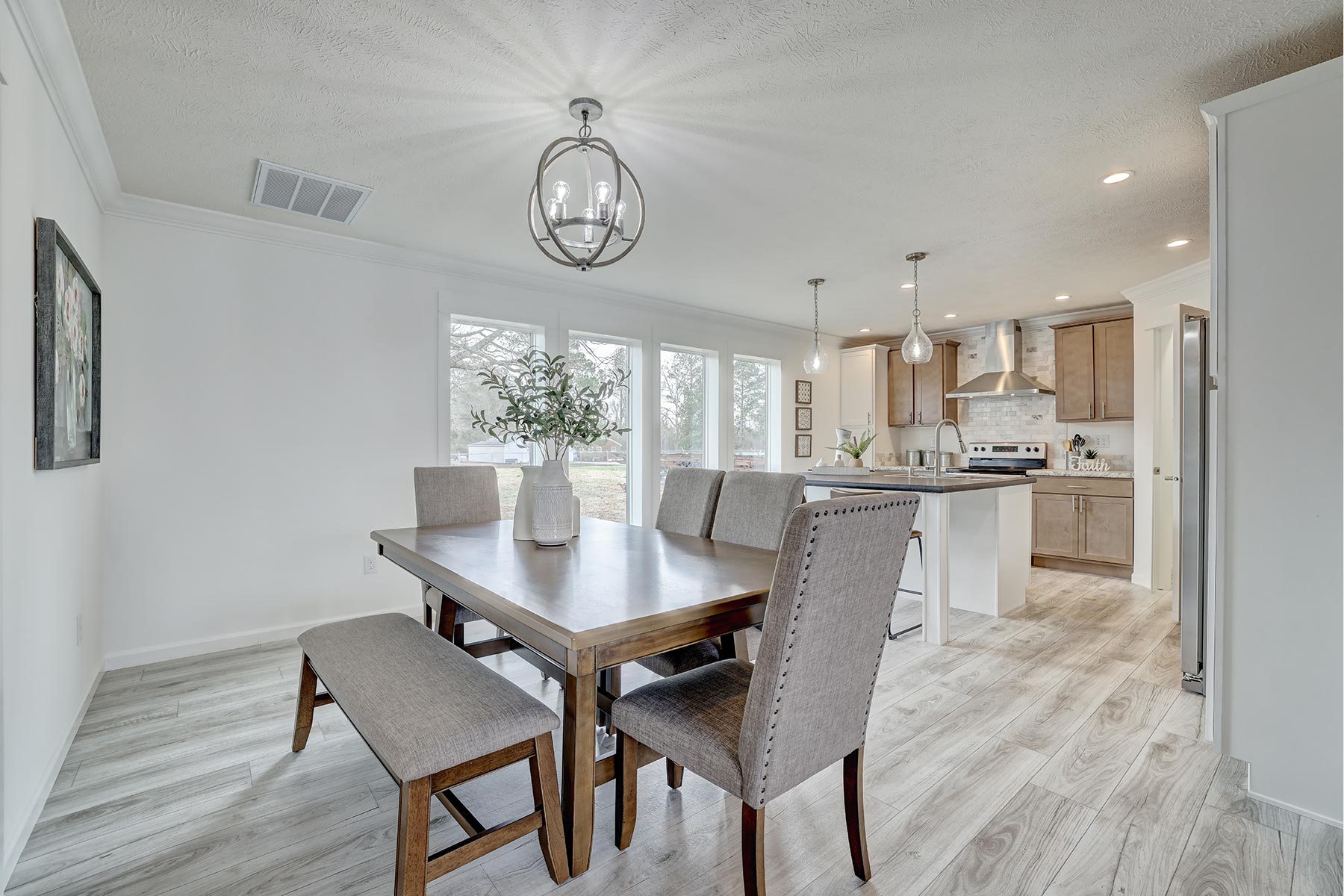 Modern dining room and kitchen with light wood flooring and neutral tones. A wood table with gray chairs sits under chic lighting. Bright and airy ambiance.