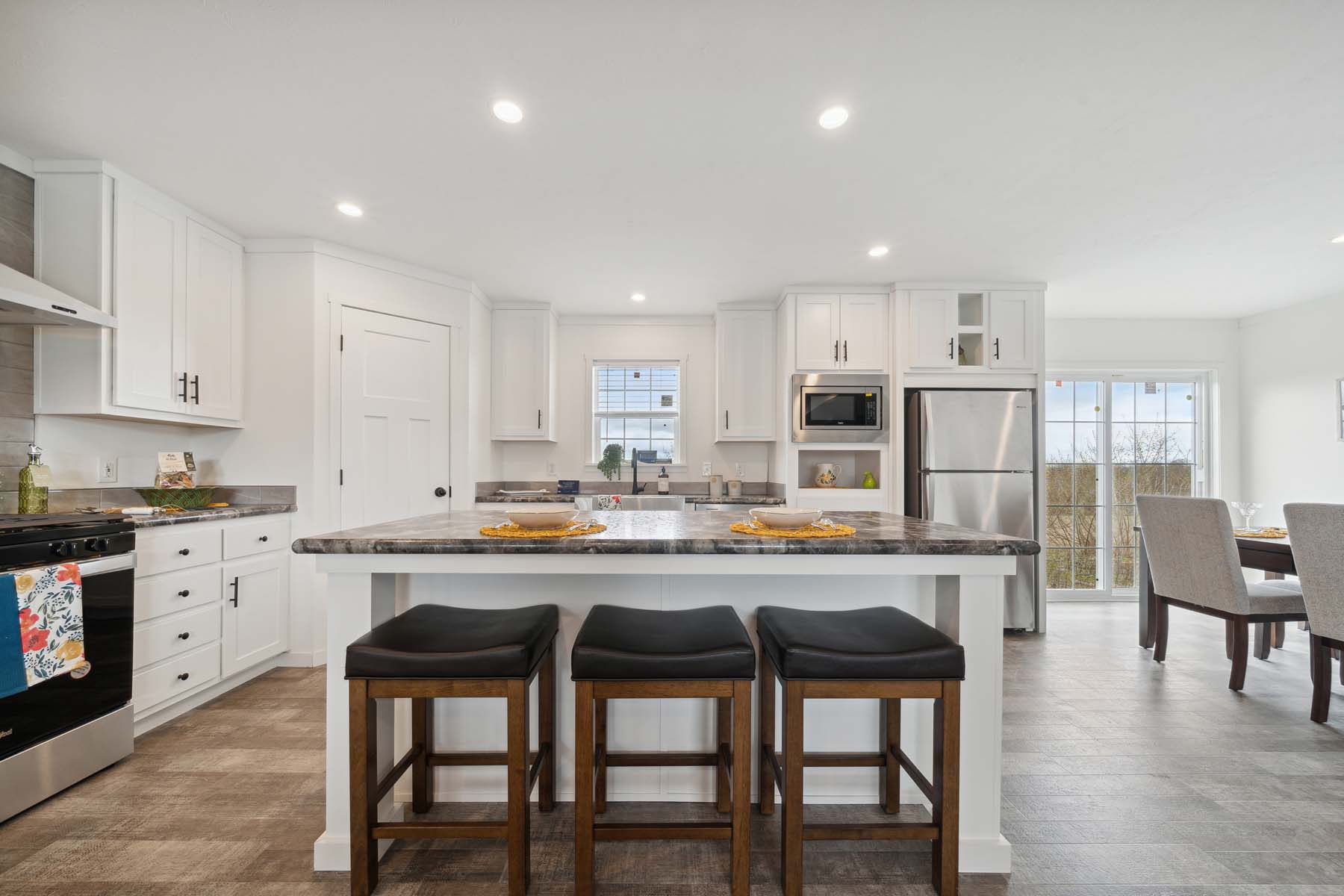 Bright kitchen with a large island, three stools, white cabinets, and stainless steel appliances. A table set is visible near glass doors on the right.