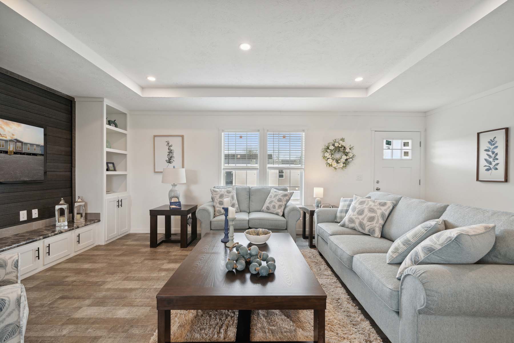A bright, cozy living room with light gray sofas, decorative pillows, and a dark wooden coffee table. Soft lighting, wall art, and a floral wreath add warmth.