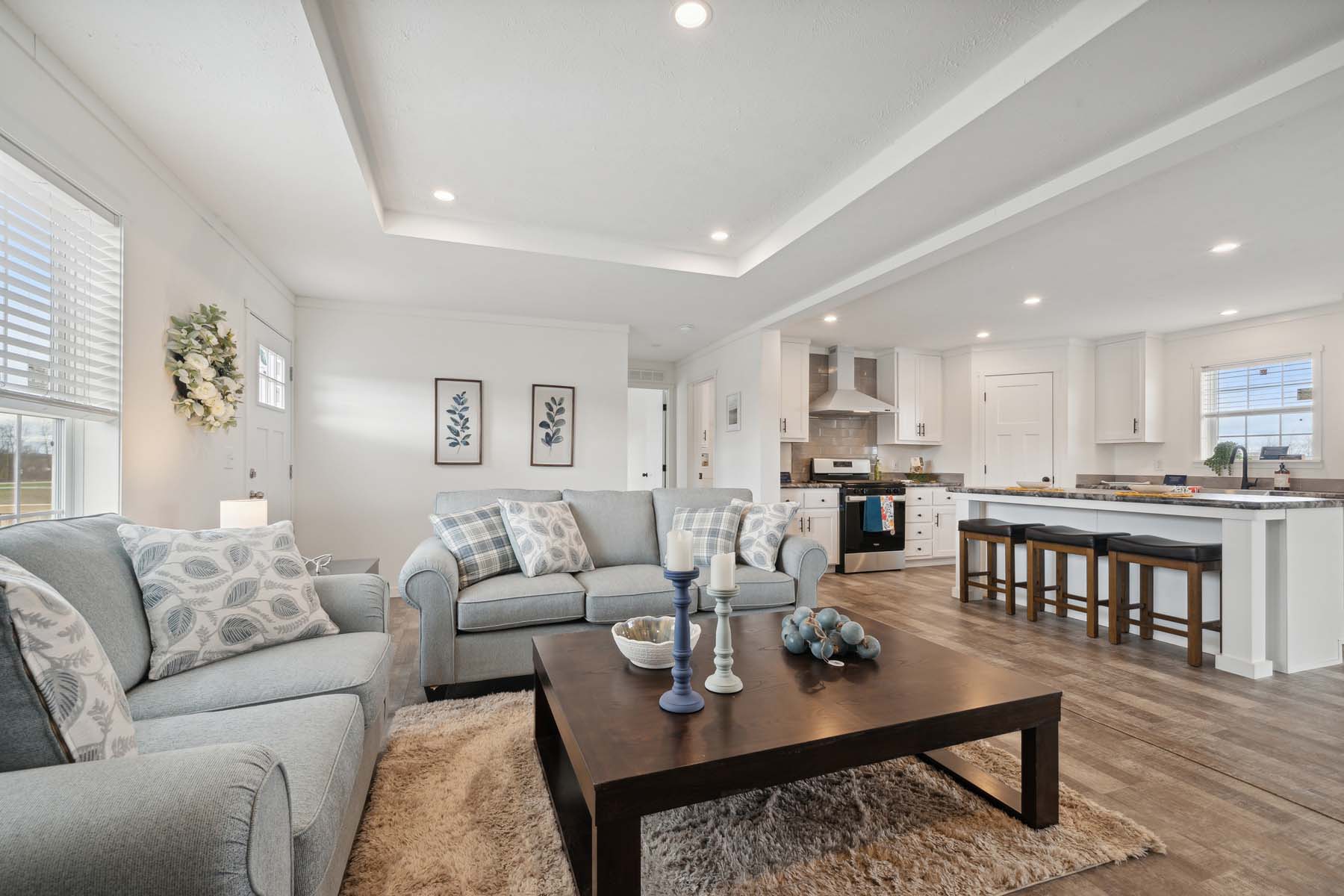 Modern living room and kitchen with open floor plan. Features gray sofas, plaid pillows, a dark wood coffee table, and island with high stools. Bright, cozy ambiance.